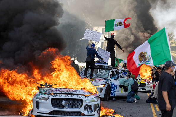 Demonstrators wave flags from atop a wrecked car, standing beside burning Waymo vehicles