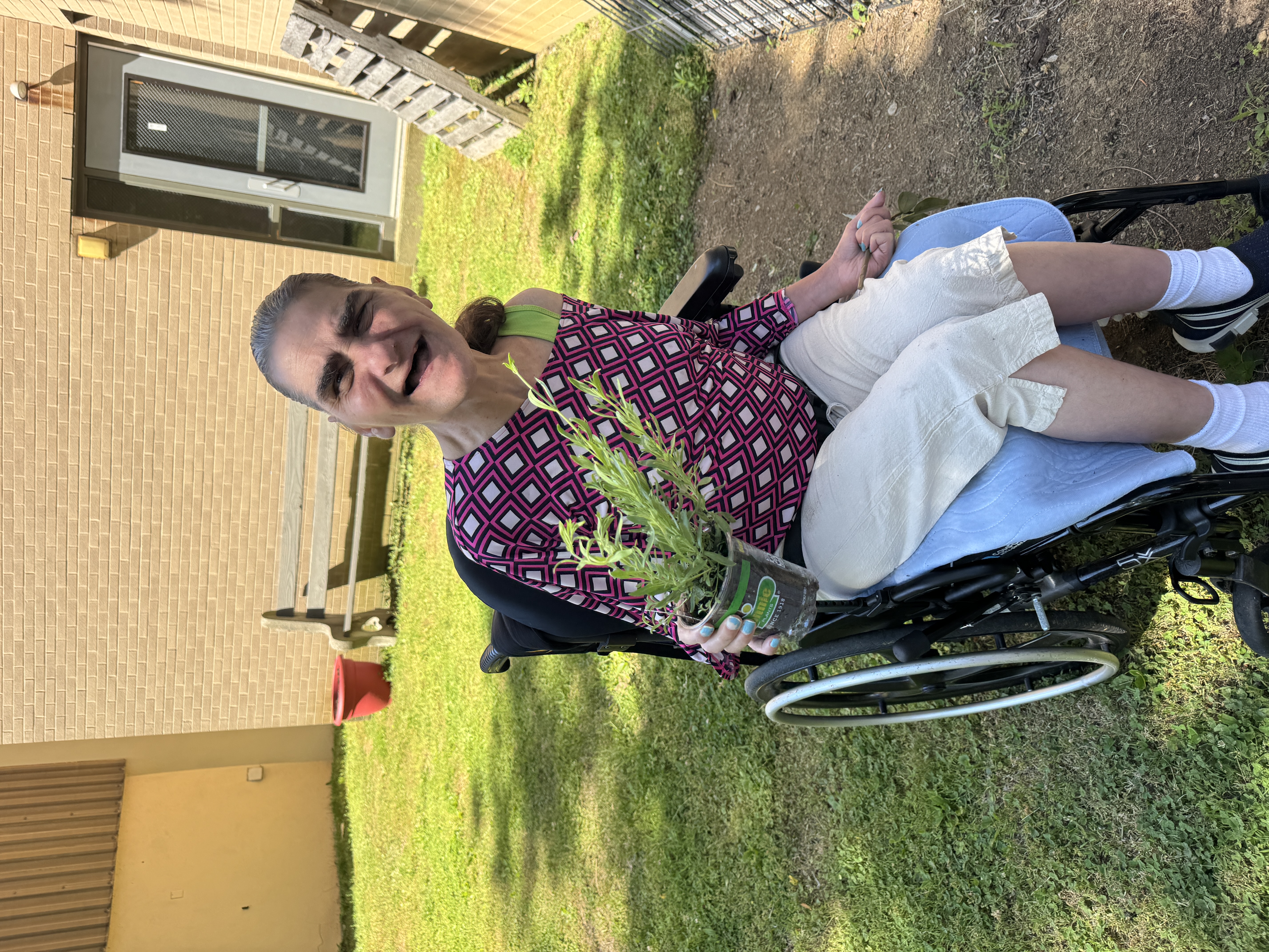 A woman is sitting in a wheelchair, she is smiling and is holding a rosemary plant, getting ready to plant it in the garden.